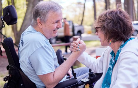 A man in a wheelchair and a woman are arm wrestling outdoors, both smiling as they celebrate Guaranty Media's 100th Anniversary with unwavering spirit and camaraderie.