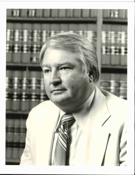 A black and white portrait features George Foster in a light-colored suit and striped tie, standing before bookshelves filled with books. The image subtly captures the legacy behind Baton Rouge Radio Station as it approaches Guaranty's 100th Anniversary.