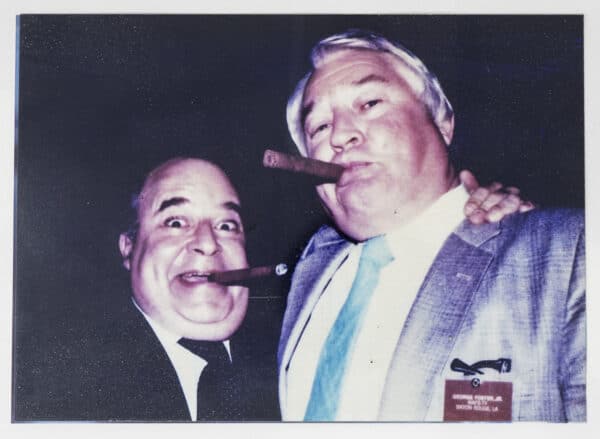 Two men in suits smile and hold cigars at a social event. One man, wearing a name badge that reads 