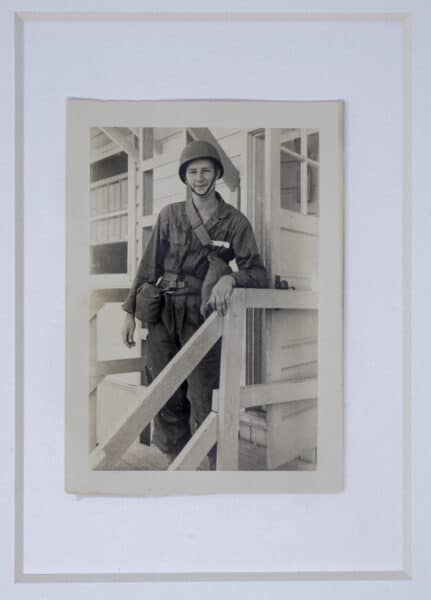 George Foster Jr, dressed in a military uniform and helmet leans on a wooden railing in front of a building, smiling at the camera.