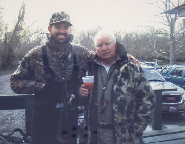 Flynn Foster and George Foster Jr., in camouflage clothing, stand outdoors, one holding a drink as they smile. Amid the trees and parked vehicles in the background, their camaraderie brings to mind the lively spirit of a Baton Rouge radio station from Guaranty Media.