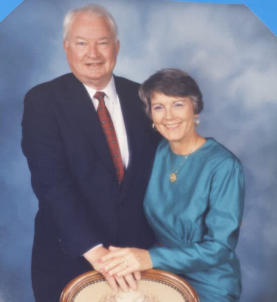 George Foster Jr. and wife, a woman in a teal dress pose together, smiling, with their hands on the back of a chair against a cloudy backdrop, celebrating Guaranty Broadcasting's 100th Anniversary.