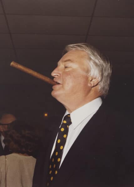 George Foster Jr. confidently holds a large cigar in his mouth, dressed in a black suit and polka dot tie. He stands indoors, embodying the timeless class of a Baton Rouge Radio Station event celebrating Guaranty Media's 100th Anniversary.