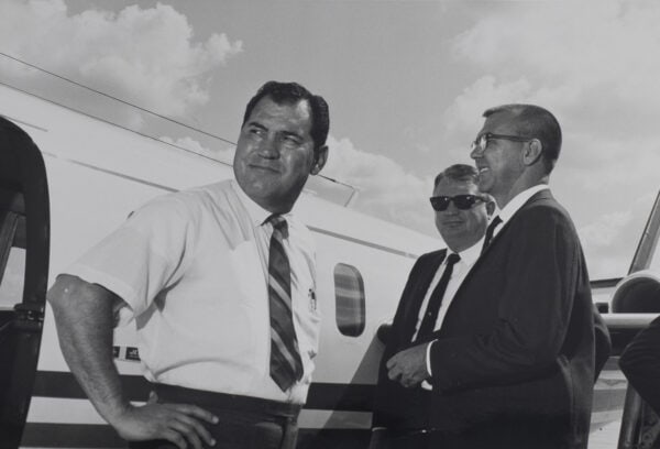 Three men in suits stand beside a small airplane. One man in a short-sleeved shirt is in the foreground, while two others, wearing sunglasses.