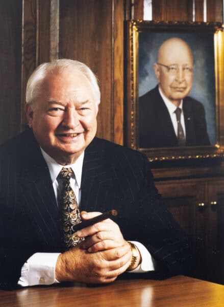 A smiling man in a suit sits at a wooden table, with a framed portrait hanging on the wooden wall behind him, celebrating Guaranty Media's 100th Anniversary.