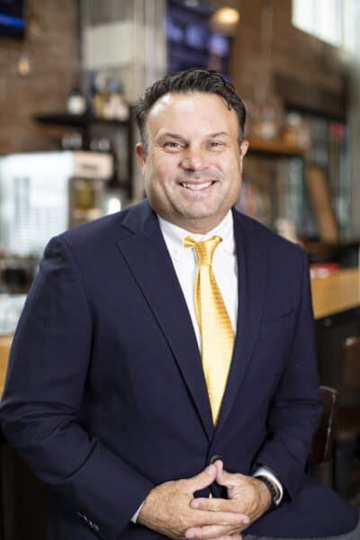 Gordy Rush in a navy suit and yellow tie is seated and smiling, exuding confidence, against a room with a brick wall and blurry background, reflecting the professionalism often associated with Guaranty Broadcasting.