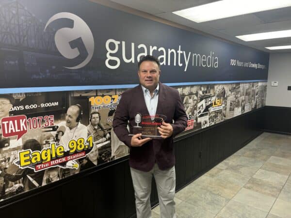 A person stands holding a plaque in front of a wall with radio station logos and 