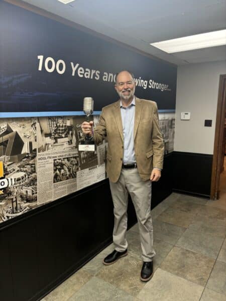 Man in a tan blazer holds a vintage-style microphone, standing in front of a wall with historical photos and text saying 