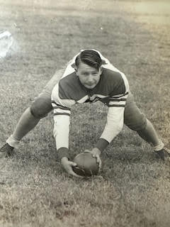 A vintage photo of a football player crouching on a field, holding a football with both hands, wearing a striped jersey and padded pants, captures the spirit reminiscent of Guaranty Foundation's enduring legacy.