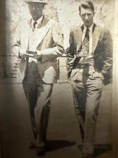 In an old, sepia-toned photograph, two men in suits and ties walk side by side. One man wears a hat and holds a book,