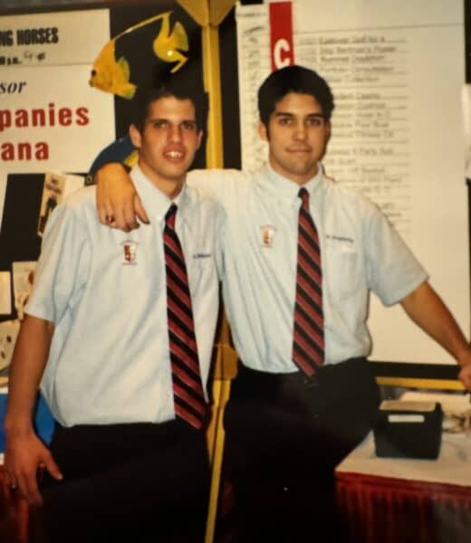 Two men in matching shirts and ties pose in front of a display board at an event.