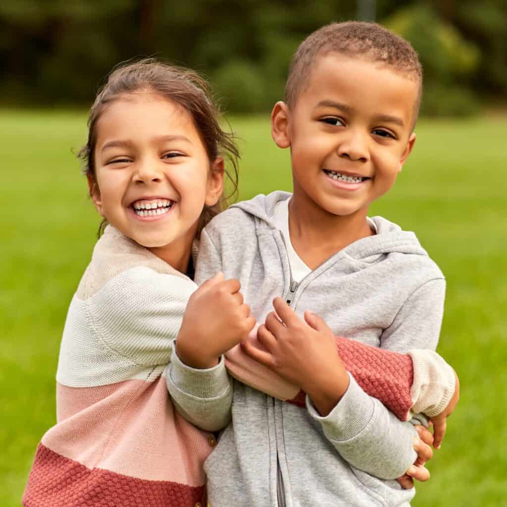 Two smiling children in casual sweaters hug outdoors
